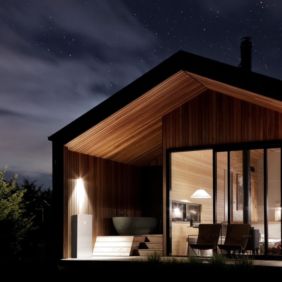 Tesla Powerwall 3 installed outside a modern wooden home, illuminated at night. The image showcases the Powerwall's sleek design and energy storage capabilities, providing backup power to the home even in off-grid settings. This setting highlights the Tesla Powerwall 3’s integration with sustainable living and home energy management solutions.